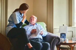 Older man sitting in a chair talking to a nurse who is sitting on the arm rest of the chair.