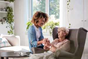 Elderly woman sitting in a comfortable chair holding hands with a young woman kneeling next to her.