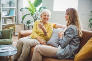 An elderly woman and a young woman sitting on a couch, drinking water, and having a conversation while smiling.