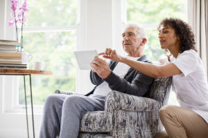 Female nurse kneeling next to an elderly man sitting in a chair. The nurse is pointing to a paper that the man is holding up in front of him.