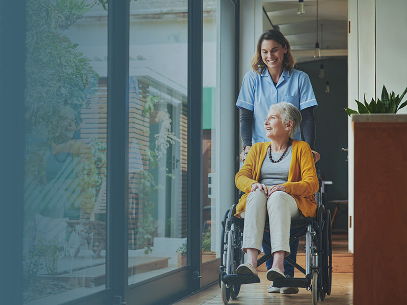 Young woman nurse pushing elderly woman in a wheelchair down a hallway next to large windows.