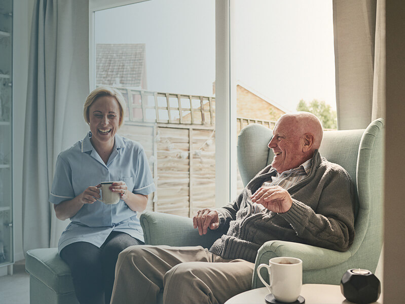 Older man sitting in a chair laughing with a young woman nurse sitting across from him with tea in her hand.