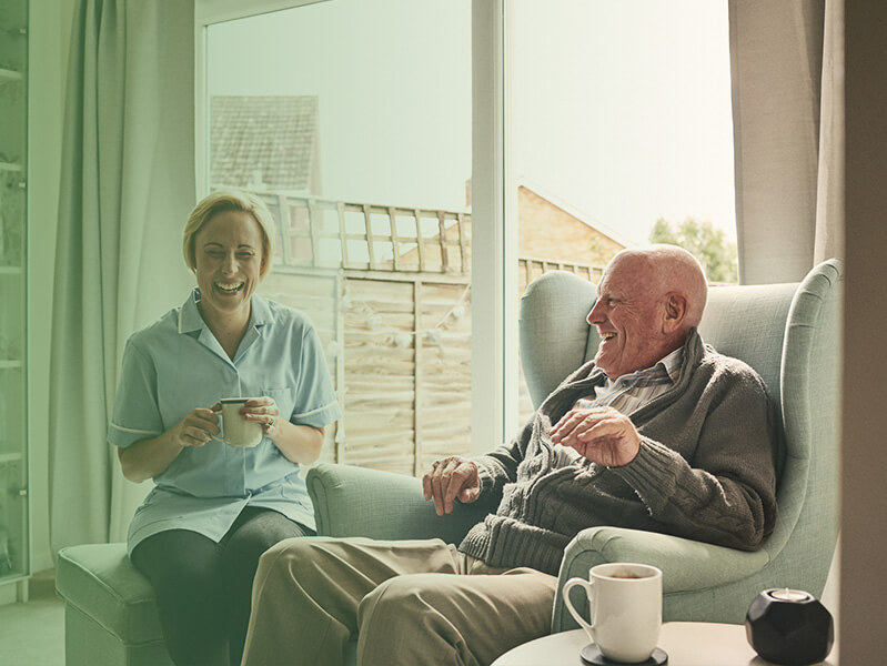 Older man sitting in a chair laughing with a woman nurse in an assisted care living facility 